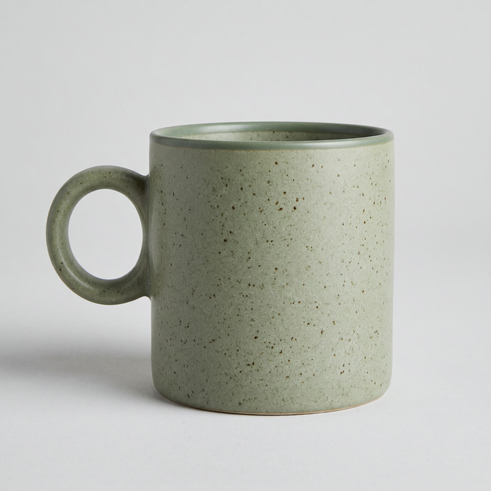 A speckled sage-green stoneware mug with a round handle on a light gray background