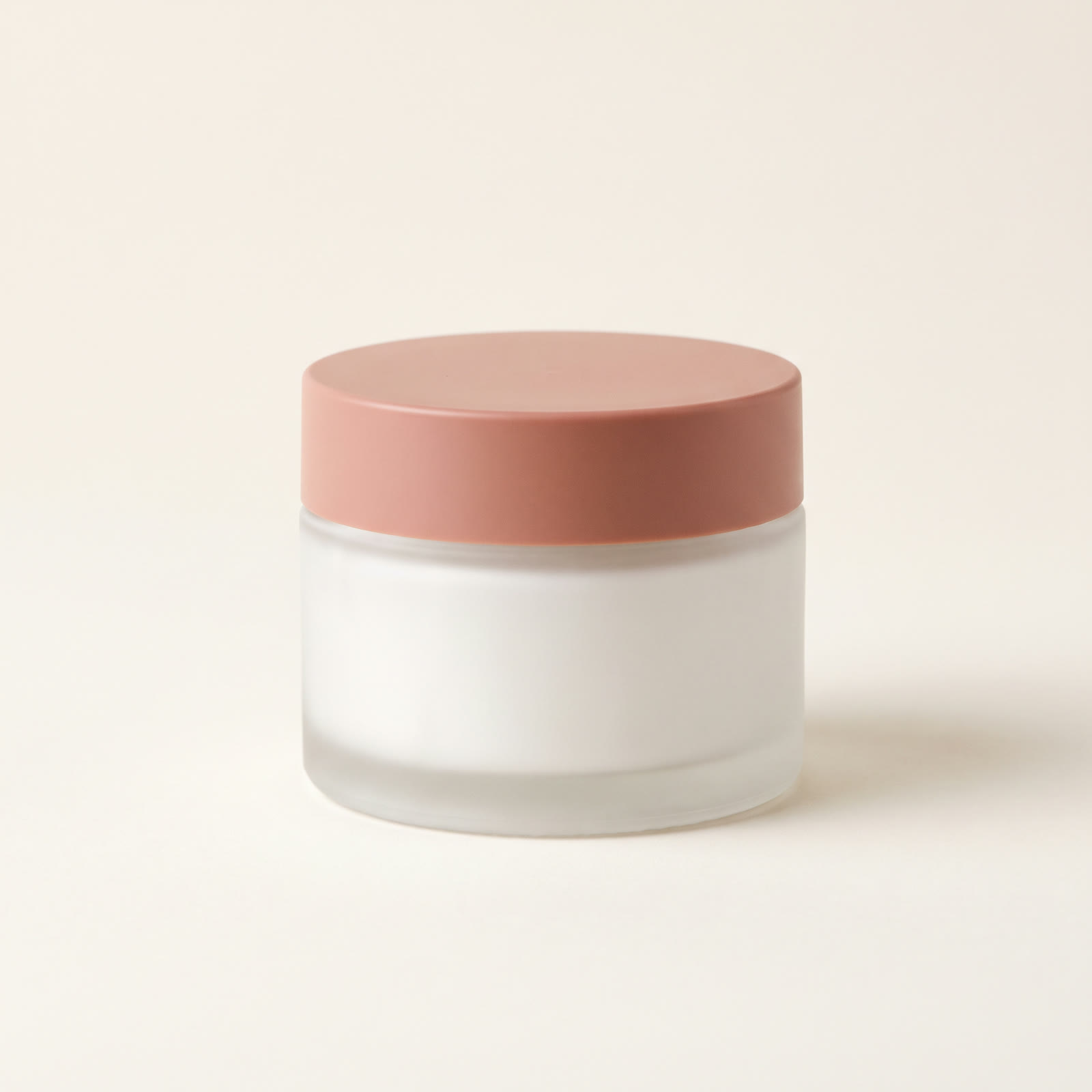 A frosted white glass jar of face cream with a matte clay-pink lid on a plain warm-white background