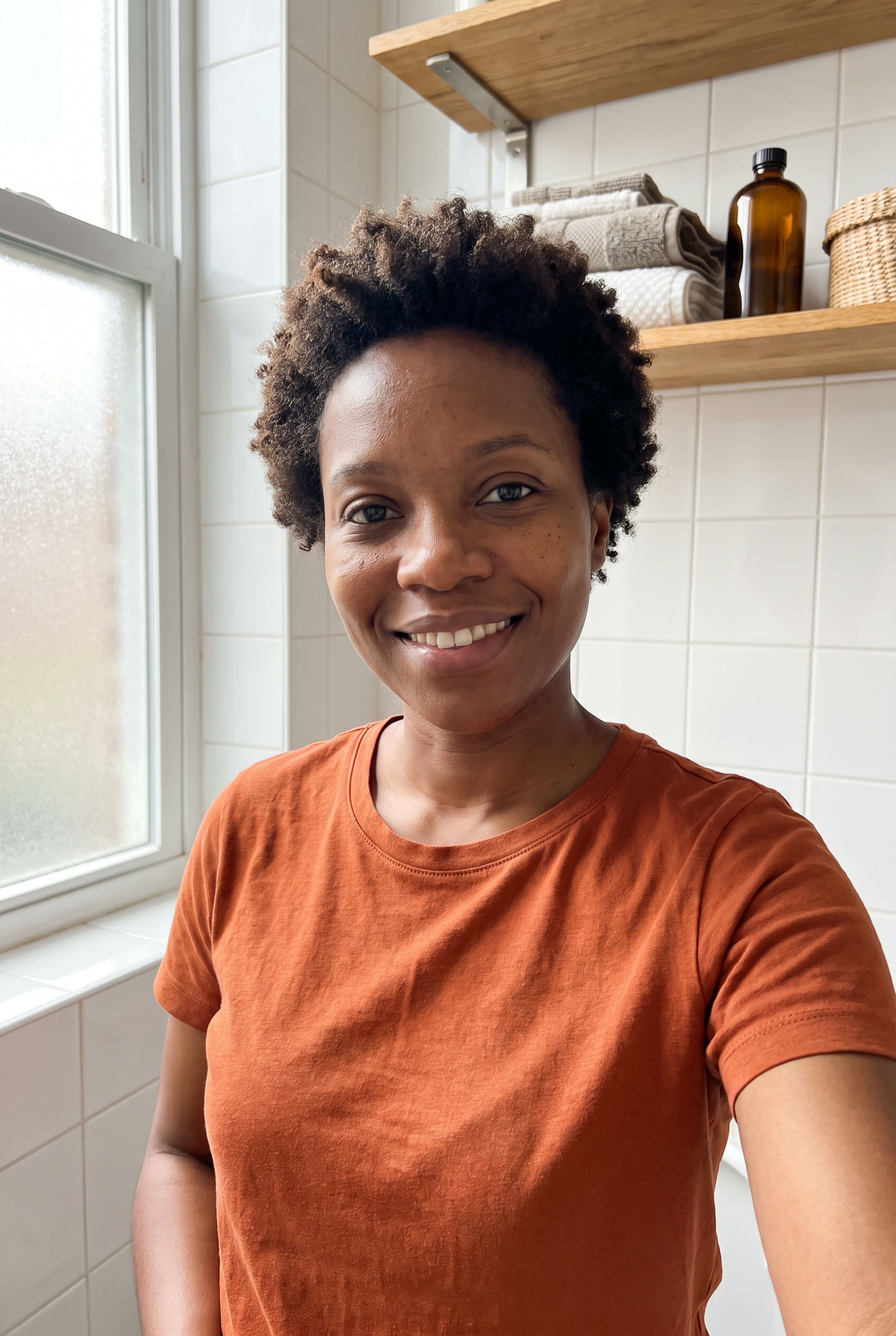 A Black woman with short natural coily hair and a rust-orange T-shirt stands in a bright tiled bathroom, framed from the waist up