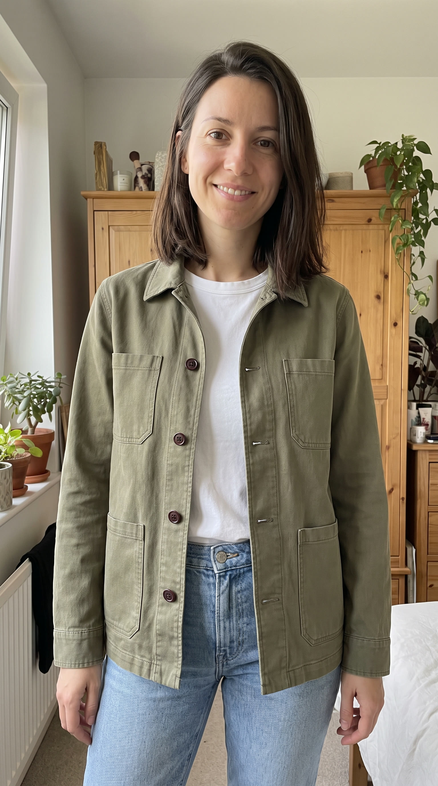 The same woman in a bedroom wearing the olive jacket open over a white T-shirt and light jeans