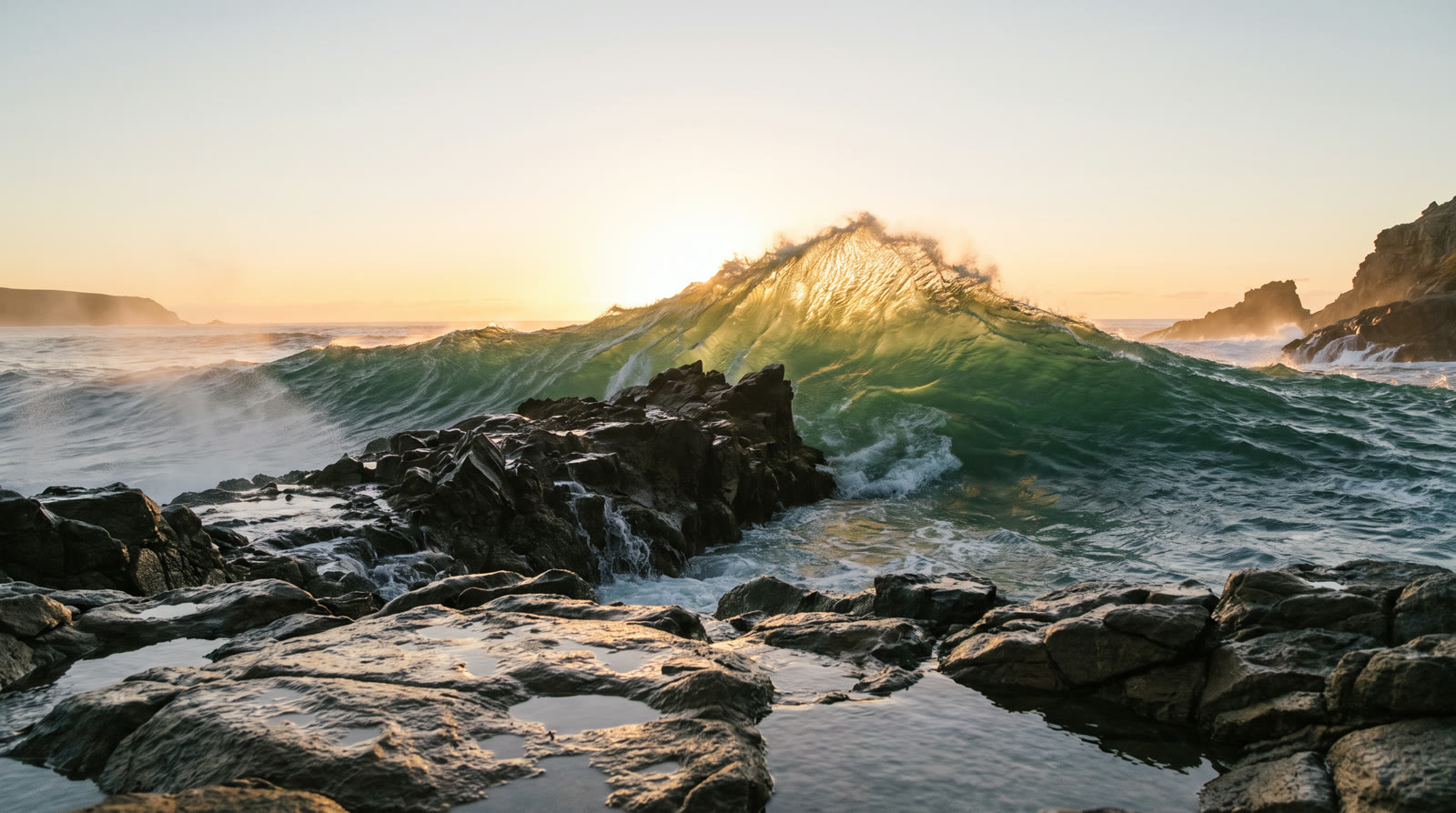 A tall green wave rising behind a dark rock outcrop at sunrise