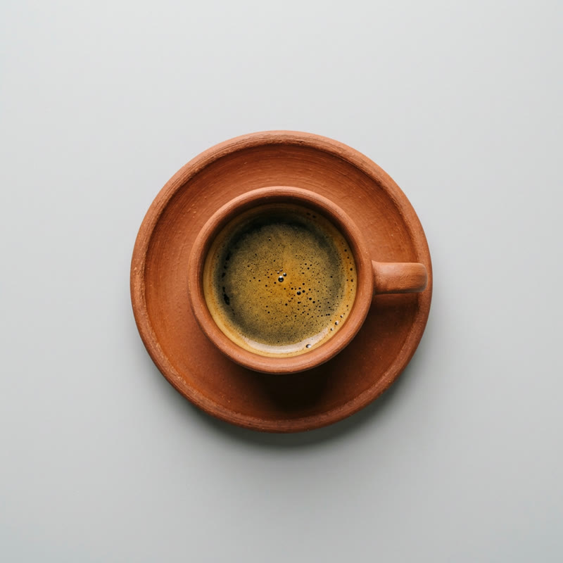 A terracotta espresso cup of coffee on its saucer on a light grey background