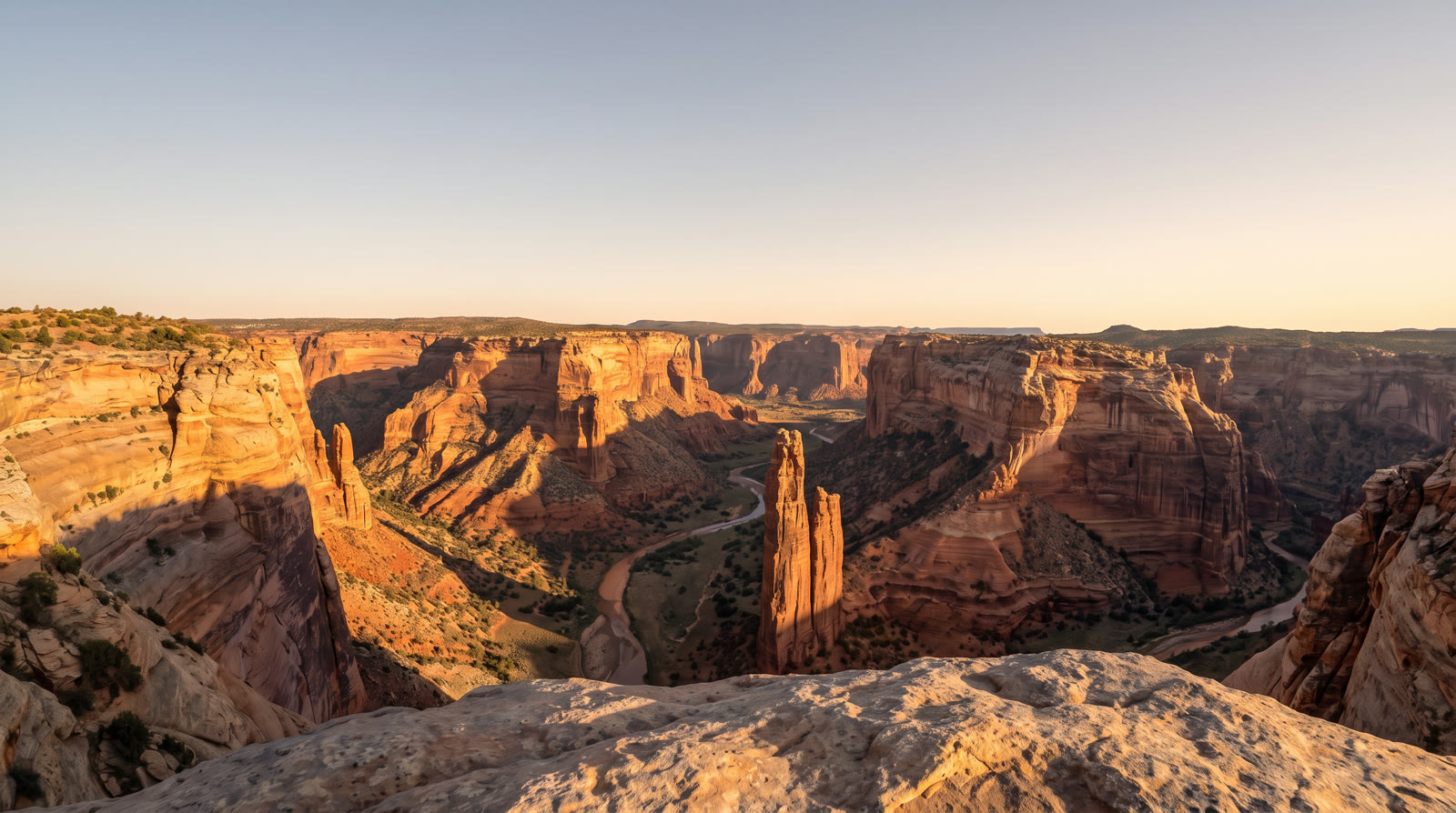 The same canyon from the same ledge at sunrise, warm light on the walls under a clear sky