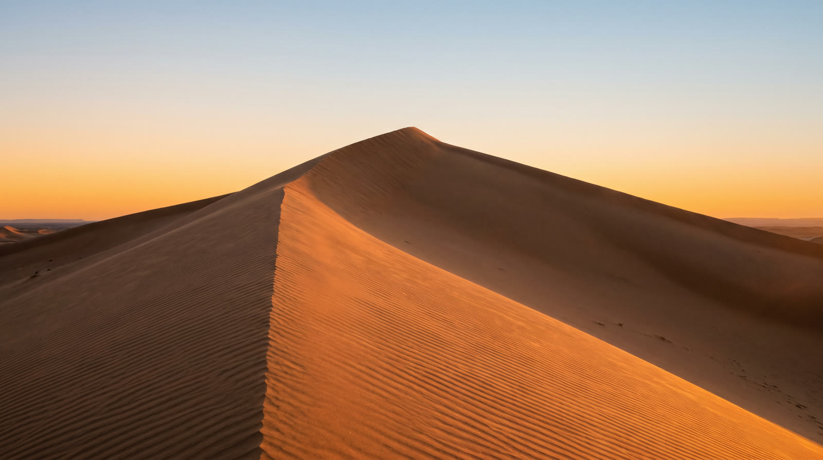 A tall sand dune at sunset, its sharp crest running across the frame and wind ripples on the near slope