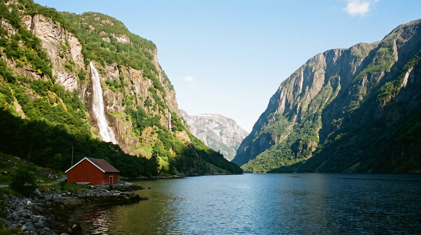 A narrow fjord with steep green cliffs, a waterfall and a red boathouse on the near shore