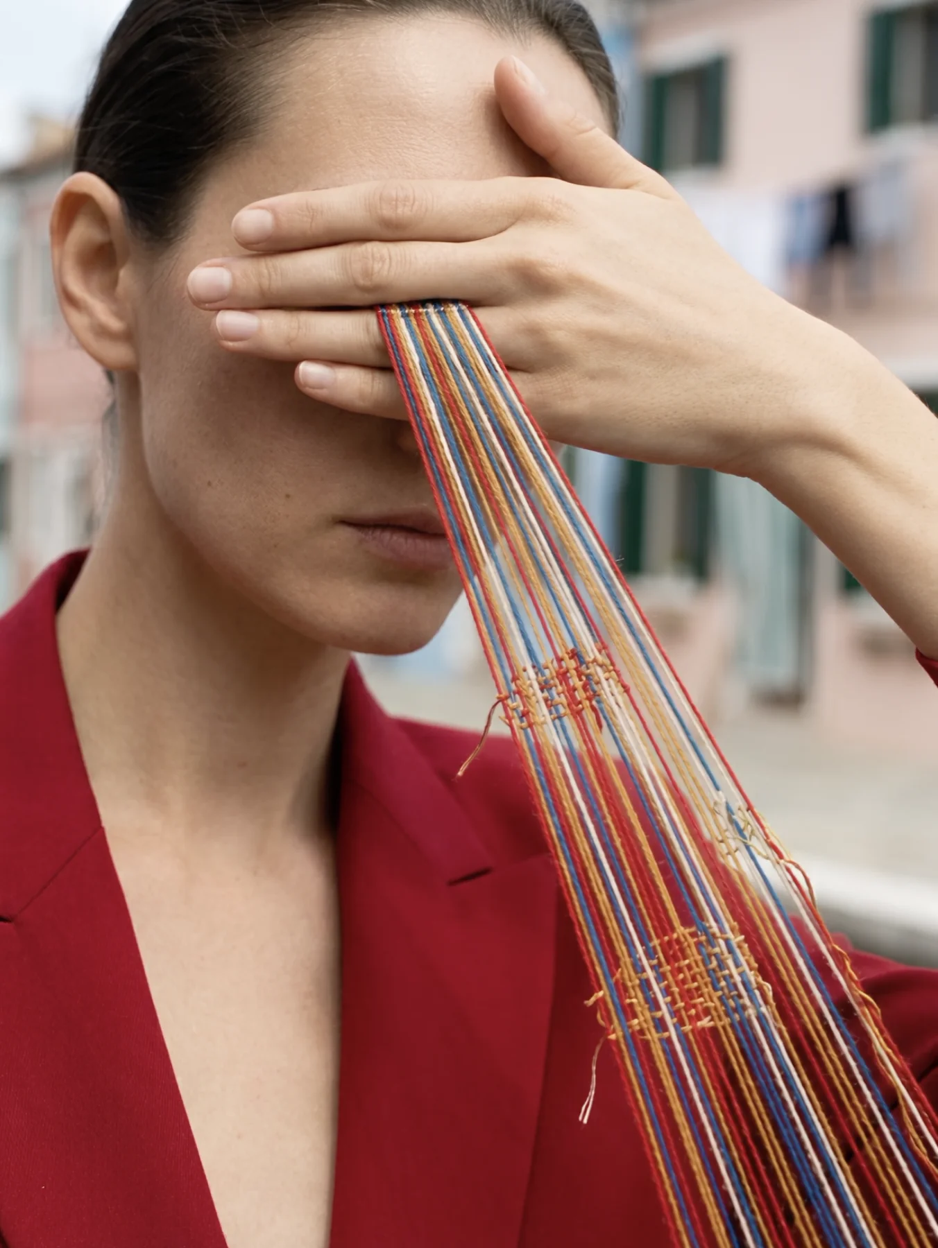 A model in a red suit covers her eyes with one hand while a flat beam of loose, multicolored stitching runs from between her fingers out of the bottom of the frame, pastel houses behind her.